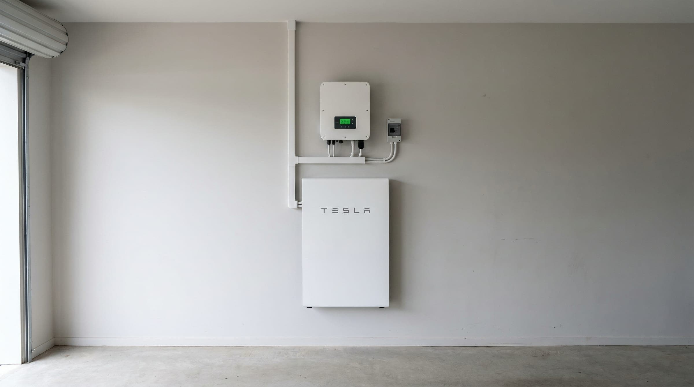 A wall-mounted Tesla Powerwall home battery and hybrid inverter installed on a clean NZ garage wall, with tidy cabling and an isolation switch.