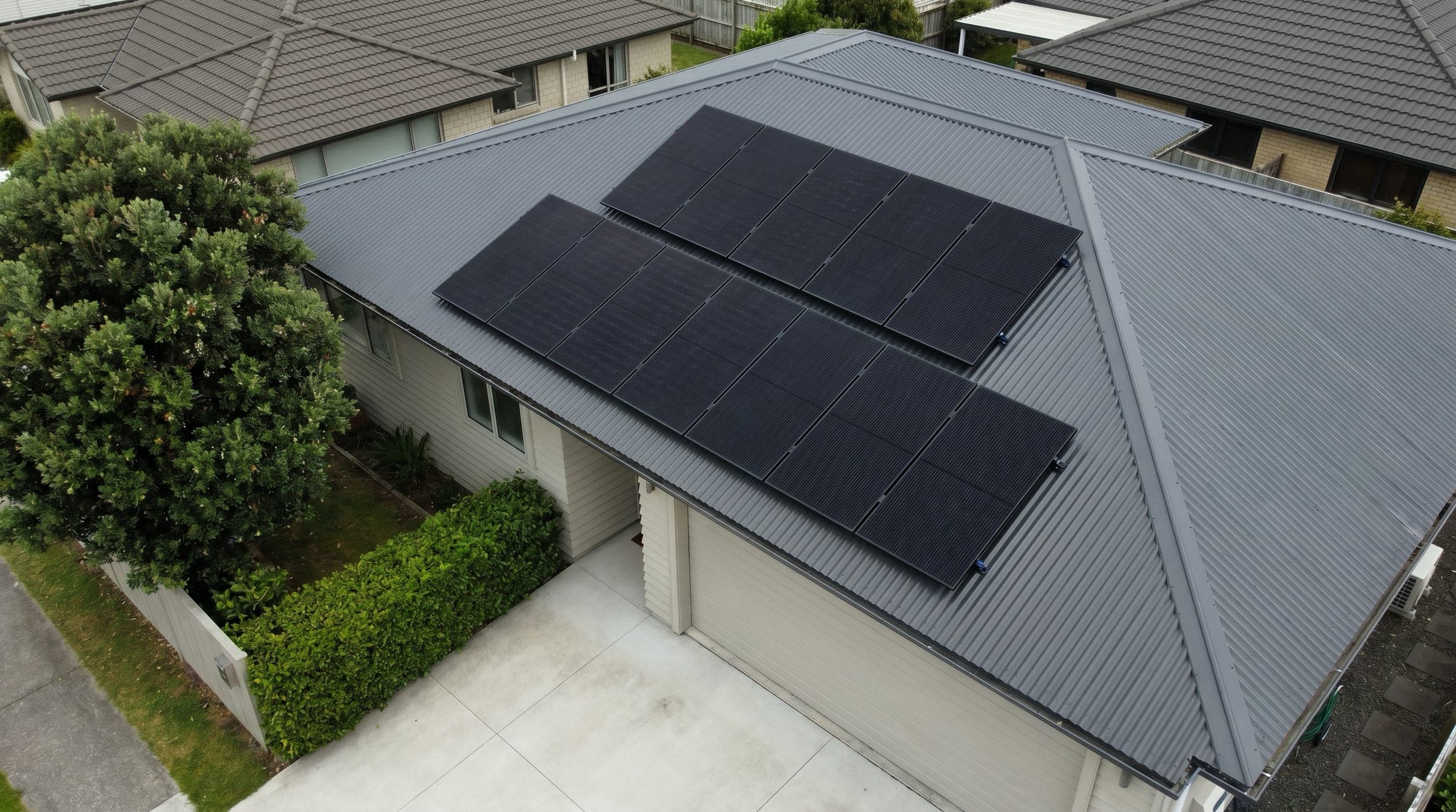 Aerial documentary photo of a typical NZ residential 6.6 kW solar install on a charcoal corrugated steel roof, with all-black monocrystalline panels laid out tidily across the north-facing pitch