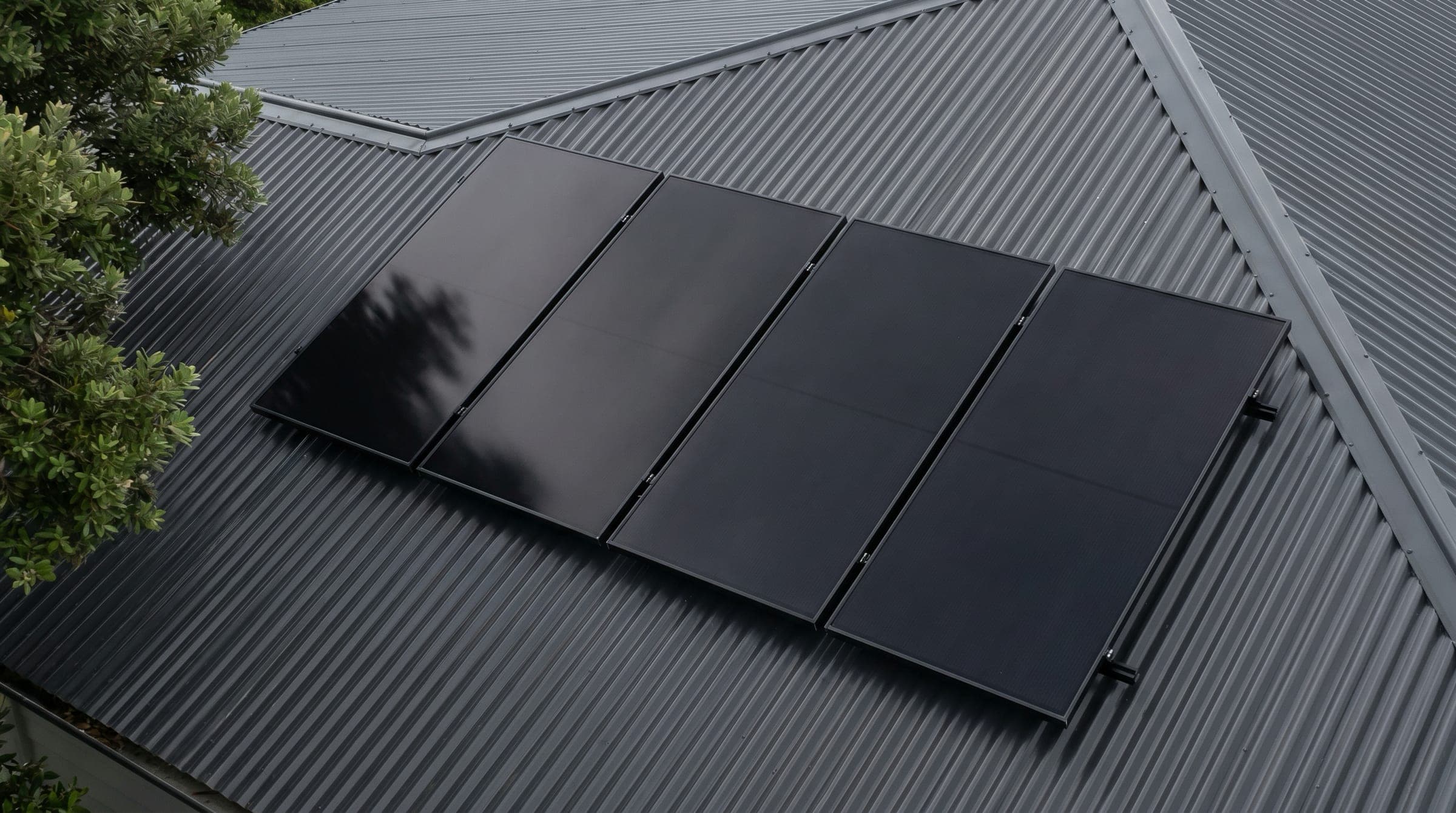 Tight documentary close-up of all-black solar panels on a charcoal corrugated steel roof with a soft diagonal tree-branch shadow creeping across the corner of one panel