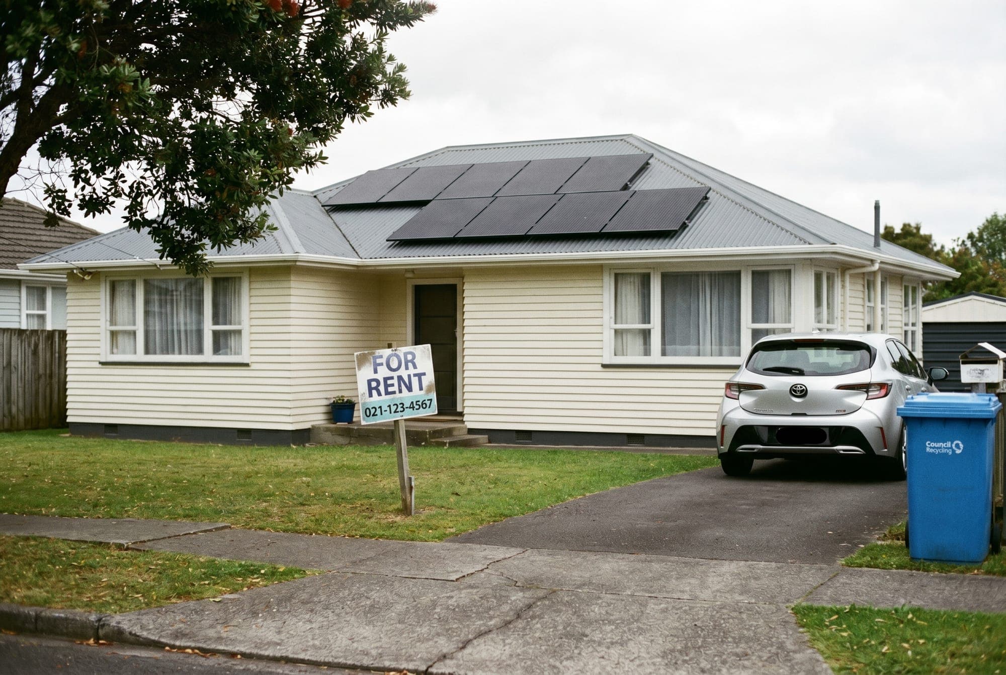A New Zealand weatherboard rental house with solar panels on the roof and a For Rent sign on the front lawn